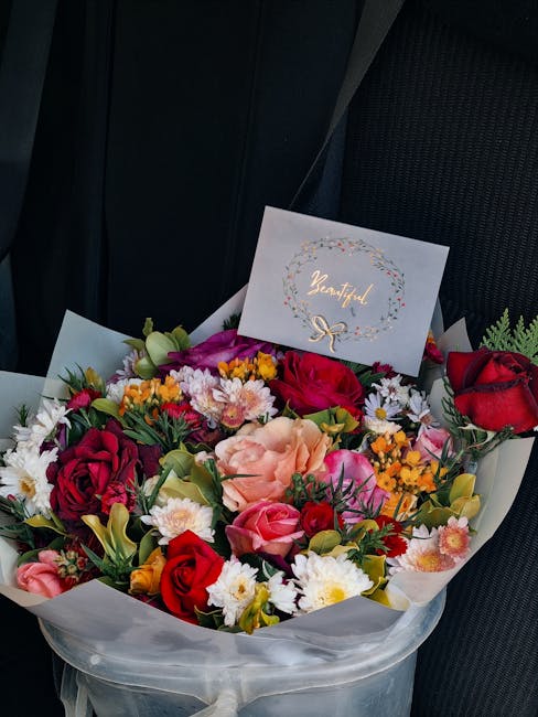 A vibrant floral bouquet arranged with a mix of red roses, peach carnations, pink spray roses, white daisies, and small yellow and orange flowers, all nestled among green foliage. The arrangement is w