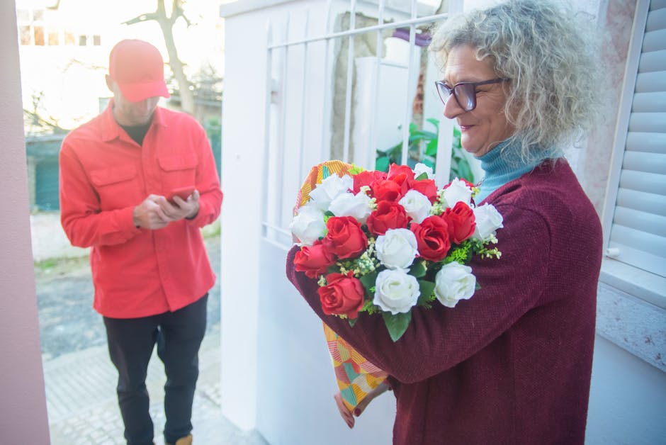 A woman with curly gray hair and glasses is holding a vibrant bouquet of fresh red and white roses, wrapped in colorful patterned paper. She is standing near a white window frame in natural daylight, 