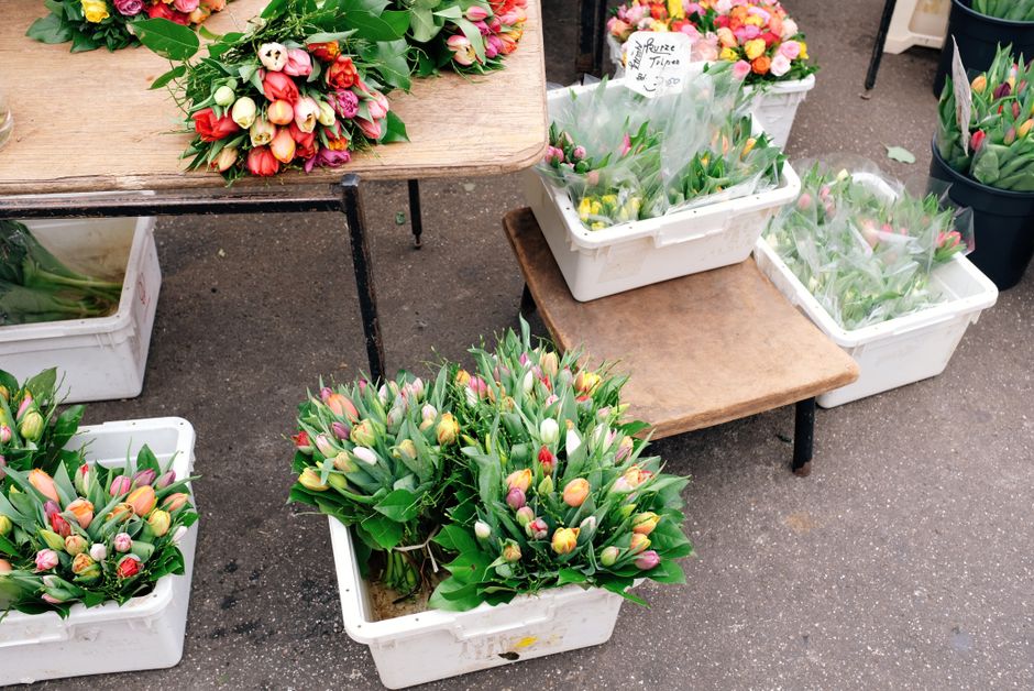 A display of fresh tulip bouquets and flower arrangements at an outdoor market stall, with an assortment of pink, white, yellow, and orange tulips arranged in white plastic crates and tied bundles, so