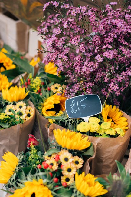 A vibrant display of fresh flower bouquets featuring bright yellow sunflowers, small white daisies, and red accents, wrapped in brown paper, arranged on a market stand. Behind the bouquets, there is a