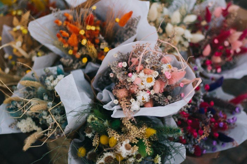 A close-up of a bouquet arranged with a mixture of small, delicate flowers including pink and white daisies, dried pink and beige grasses, and tiny pink blossoms, wrapped in white tissue paper. The fl