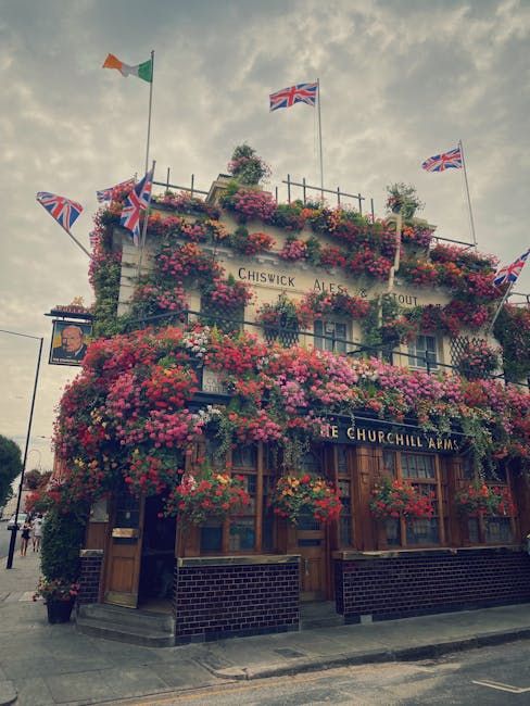 The exterior of the Churchill Arms pub in Pimlico is extensively decorated with vibrant pink and red azalea and geranium flowers covering the façade, creating a lush floral display. Union Jack flags 