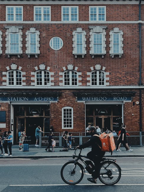 The image depicts the exterior of Waterloo Station on Waterloo Road, a historic brick building with multiple evenly spaced windows, some with decorative white trims. In front of the station, a diverse