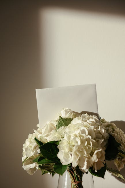 A bouquet of white hydrangeas and roses arranged with dark green leaves, placed in a clear glass vase. The flowers are fresh, with delicate petals and sturdy stems, positioned on a surface near a wall