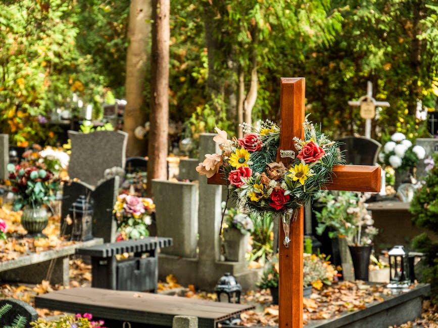 A wooden wooden cross adorned with a floral arrangement of red roses, yellow daisies, and white lilies, decorated with greenery, is positioned among a variety of headstones and grave markers at a ceme
